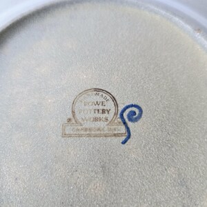 K&ouml;nnte beinhalten: Nahaufnahme eines Keramiktellers mit hellgrauer Oberfl&auml;che. Der Teller hat einen runden Stempel mit der Aufschrift "HAND MADE ROWE POTTERY WORKS CAMBRIDGE, MASS." Neben dem Stempel befindet sich ein blaues Wirbeldesign.