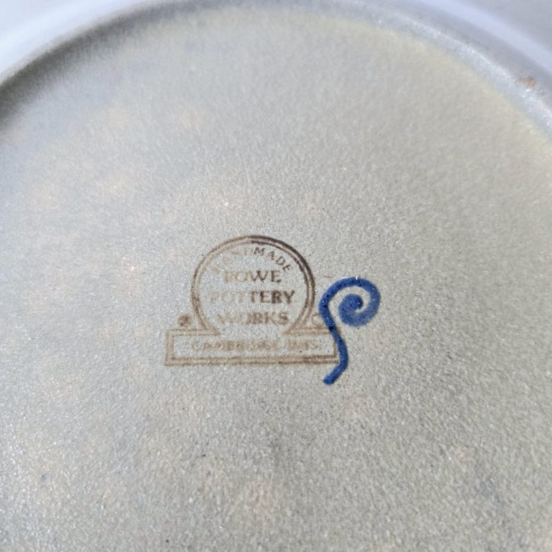 K&ouml;nnte beinhalten: Nahaufnahme eines Keramiktellers mit hellgrauer Oberfl&auml;che. Der Teller hat einen runden Stempel mit der Aufschrift "HAND MADE ROWE POTTERY WORKS CAMBRIDGE, MASS." Neben dem Stempel befindet sich ein blaues Wirbeldesign.