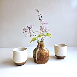 May include: A ceramic vase with a brown and white drip glaze containing light purple flowers, flanked by two matching ceramic cups featuring a white upper section and a brown base with horizontal ridges on the exterior surface of the cups.
