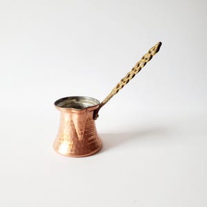 May include: A copper-colored coffee pot with a hammered texture and a brass handle. The handle has a decorative, twisted design. The pot has a wide base that tapers to a narrower opening. The interior of the pot is a darker color.