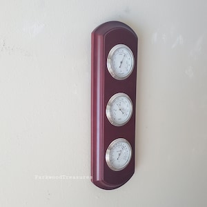May include: A mahogany-colored wooden wall-mounted weather station with three silver-colored analog gauges. The gauges measure temperature, humidity, and barometric pressure.