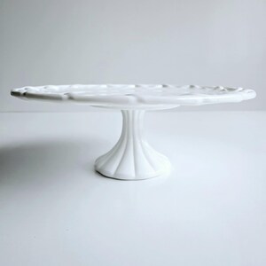 May include: White ceramic cake stand with a scalloped edge and a fluted pedestal base.