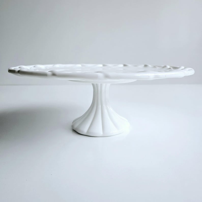 May include: White ceramic cake stand with a scalloped edge and a fluted pedestal base.