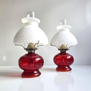 May include: Two vintage red glass oil lamps with white hobnail shades. The lamps have a round base and a clear glass chimney. The shades are a classic design, and the lamps are likely from the early 20th century. The lamps are a beautiful decorative item.