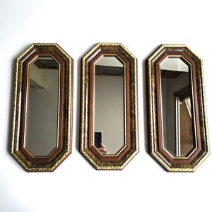 May include: Three rectangular mirrors with octagonal frames. The frames have a dark brown interior, a gold trim, and a textured, dark brown and gold outer edge. The mirrors are displayed against a white wall.