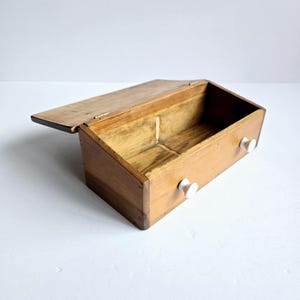 May include: A rectangular wooden box with a hinged lid and an open interior. The exterior features a natural light brown wood grain finish and two small, round white knobs attached to the front panel, providing a simple and functional design.