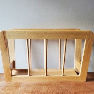 May include: A wooden magazine rack with a light brown finish and five vertical dividers. The rack has a rectangular shape and a slightly angled design.