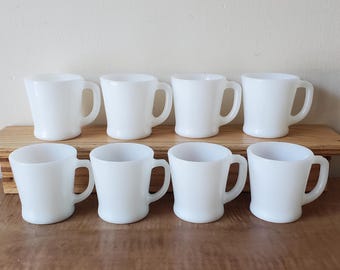May include: Eight white ceramic mugs with handles. The mugs are arranged in two rows of four on a wooden surface.