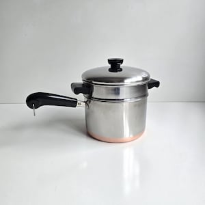 May include: A stainless steel double boiler pot with a copper-colored base. The pot features a long black handle with a metal hanging loop, two black side handles, and a matching stainless steel lid with a black knob on top for easy lifting.