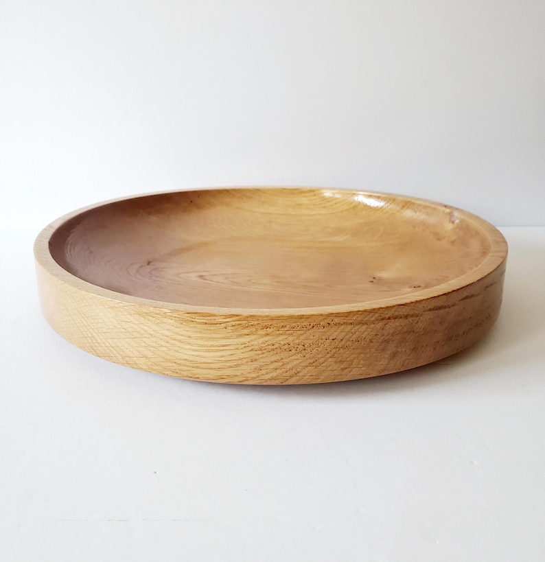 K&ouml;nnte beinhalten: Eine runde Holzschale mit hellbraunem Finish. Die Schale hat einen flachen, gew&ouml;lbten Innenraum und einen dicken, abgerundeten Rand. Die Holzmaserung ist sichtbar und verleiht dem St&uuml;ck Textur. Die Schale wird wahrscheinlich zum Servieren von Speisen oder als Dekorationsartikel verwendet. Durchmesser ca. 30 cm.