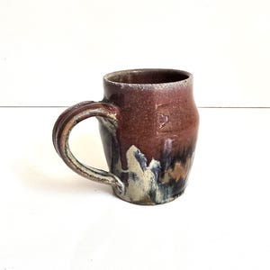 May include: A ceramic mug with a rounded body and a curved handle. The upper portion features a deep reddish-brown glaze, while the lower section transitions into a mottled pattern of cream, dark blue, and black tones with a glossy finish.