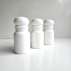 May include: Three identical white apothecary jars with rounded lids. Each jar features a cylindrical body with a narrow neck and a smooth, glossy finish. The containers are arranged in a staggered formation, showcasing their uniform design and opaque white color.
