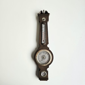 May include: A dark brown wooden barometer with a thermometer and two gauges. The barometer has a circular gauge with a gold-colored rim and a needle pointing to the current pressure. The thermometer displays the temperature in Fahrenheit.