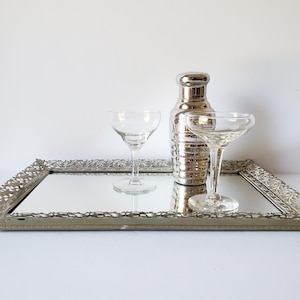 May include: A vintage mirrored tray with a decorative silver frame holds two clear stemmed cocktail glasses and a silver cocktail shaker. The tray reflects the objects, creating a classic barware display.