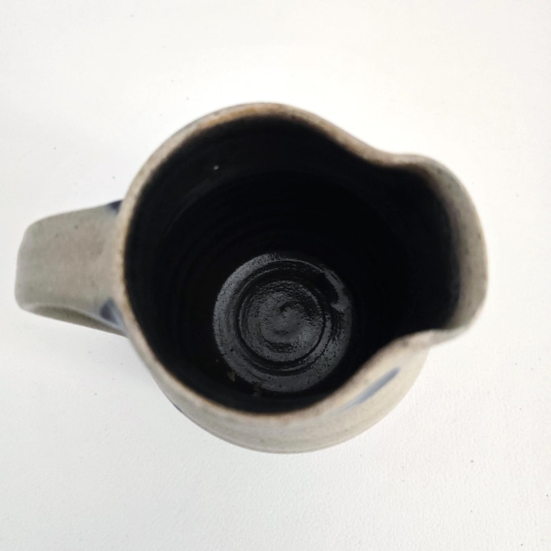 Op de afbeelding: Een handgemaakte keramische kan met een handvat. De kan is lichtgrijs met donkerblauwe accenten en een donker interieur. De kan heeft een tuit om gemakkelijk te schenken en is geschikt voor het serveren van dranken.