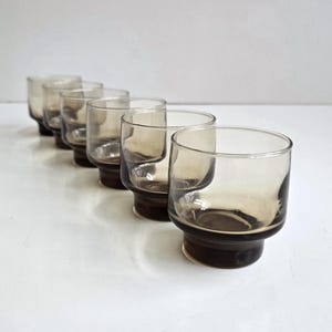 May include: A set of six small, round drinking glasses arranged in a diagonal line. Each glass is made of translucent, smoke-colored glass with a darker, thicker base that tapers slightly toward the bottom, featuring a smooth, cylindrical rim.