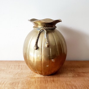 May include: A golden-colored, round vase with a ruffled top and vertical grooves. A rope-like detail with two decorative balls hangs from the top. The vase sits on a wooden surface.