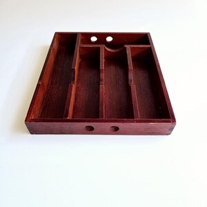 May include: A dark reddish-brown wooden cutlery tray, rectangular in shape, with multiple compartments. The tray has a long, narrow section and several smaller, square sections. Two small holes are drilled into the front. The wood has a rich, polished finish.