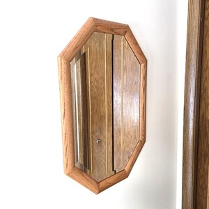 May include: An octagonal wooden mirror with a light brown frame and a vertical wood-paneled center. The mirror is mounted on a white wall. The frame has a beveled edge.
