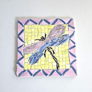 May include: A square mosaic tile featuring a dragonfly design. The dragonfly has blue and purple wings, black body, and is set against a yellow background. The tile is framed with pink and blue geometric shapes.