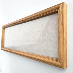 May include: A rectangular picture frame with a light brown wooden border and a textured, off-white interior surface. The frame is viewed from an angled perspective, highlighting the grain of the wood and the depth of the frame's edge.