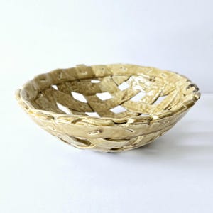 May include: A round, beige ceramic bowl featuring a woven basket-style design with open lattice gaps throughout the body. The surface has a glossy, glazed finish with visible texture and a slightly uneven, hand-formed rim and structure.