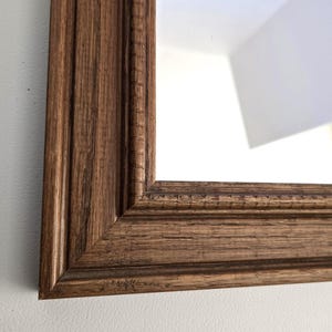 May include: Close-up of a rectangular mirror with a brown wooden frame. The frame has a detailed, textured design with vertical and horizontal grooves. The mirror reflects a white wall and a corner.