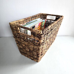 May include: A rectangular woven basket made of dark brown natural fibers featuring an open-top design with integrated side handles. The basket is filled with several magazines and papers, showcasing a textured, braided construction throughout.