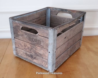 Op de afbeelding: Vintage houten melkkrat met metalen hoekverstevigingen. De krat heeft de woorden "ARNOLD DAIRIES" en "DEPOSIT CASE" erop. Het heeft handgrepen en is gemaakt van verweerd hout.
