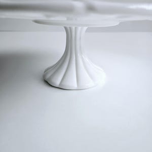 May include: White ceramic cake stand with a fluted base. The stand is round and has a smooth, glossy finish.