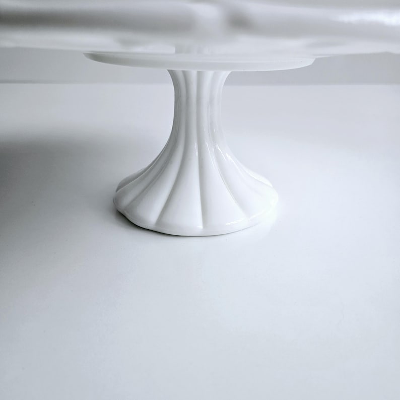May include: White ceramic cake stand with a fluted base. The stand is round and has a smooth, glossy finish.