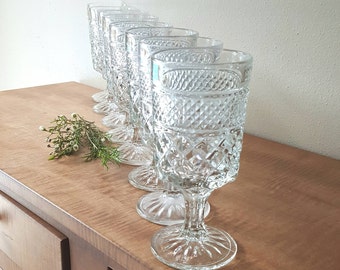 May include: A set of eight clear glass goblets with a diamond pattern. The goblets are arranged in a row on a wooden surface.