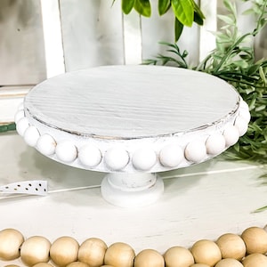 May include: White wooden cake stand with a beaded edge. The stand is on a white wooden surface with a strand of wooden beads in the foreground.