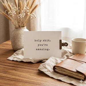 May include: A rectangular white card featuring the black typewriter-style text holy shit. you're amazing. The card is held by a bronze-colored metal clip and rests on a piece of light-colored fabric next to a brown leather journal and a white mug.