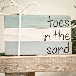 May include: A stack of three wooden blocks painted in shades of blue and white. The blocks are tied together with twine and have the words "toes in the sand" written in black paint.