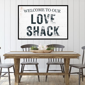 Pode incluir: Uma placa de madeira rústica com tinta branca e letras pretas que diz "Bem-vindo ao nosso Love Shack". A placa está pendurada em uma parede branca em uma sala de jantar com uma mesa de madeira e quatro cadeiras cinzas.