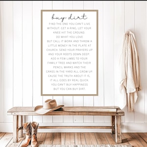 Op de afbeelding: Een beige bord met de tekst "buy dirt" en een citaat over het belang van hard werken en familie. Het bord hangt aan een witte muur boven een houten bank met een bruine cowboyhoed erop.