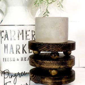 May include: A set of four wooden plant stand risers with a dark brown stain. The risers have a round shape and are stacked on top of each other. Each riser has a row of small wooden balls around the edge. A gray planter with a green plant is sitting on top of the risers.