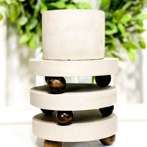 May include: A set of three tiered, round, wooden candle holders with a light gray finish. Each tier has a set of wooden ball feet and is stacked on top of each other. The top tier has a white ceramic cylinder on top.