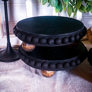 May include: Two black wooden tiered cake stands with a beaded trim and wooden feet. The stands are stacked on top of each other.