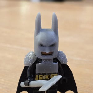 May include: A Lego Batman figurine with a gray cowl and a black cape. The figure is holding a white blade. The chest piece has a gold belt design. The figure has gray shoulder pads.