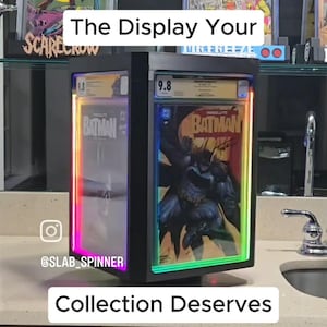 May include: A black, rectangular, rotating display case for graded comic books, featuring integrated LED lighting strips along the inner edges. The case holds a slabbed Absolute Batman comic book with a 9.8 grade label visible at the top.