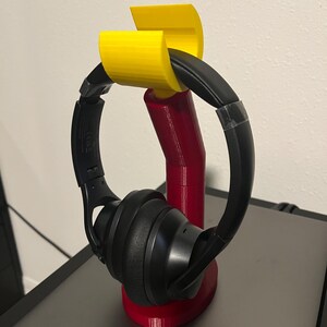May include: A red and yellow headphone stand holding a pair of black headphones. The stand is made of plastic and has a curved design to cradle the headphones. The stand is on a dark surface.