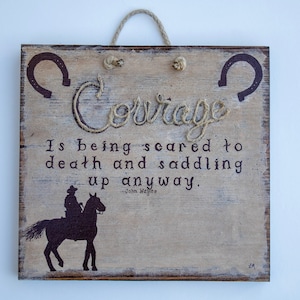 May include: Wooden sign with a rustic design. The word "Courage" is spelled out in rope. The sign features a quote by John Wayne and a silhouette of a cowboy on a horse, with two horseshoe designs.