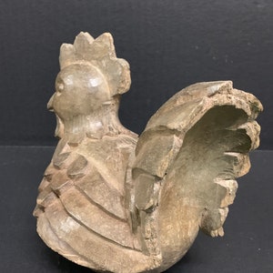 Puede incluir: Una figurita de gallo de madera tallada a mano con una cresta y plumas de cola detalladas. El gallo est&aacute; pintado de color marr&oacute;n claro y tiene un aspecto r&uacute;stico y antiguo.