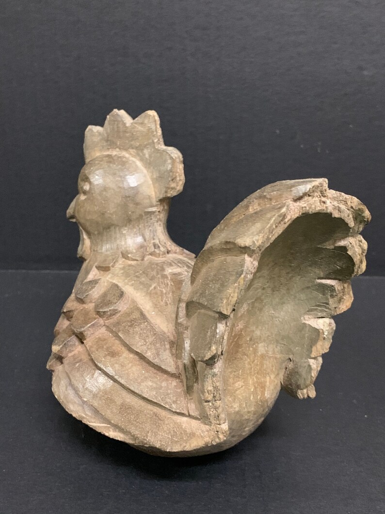 Puede incluir: Una figurita de gallo de madera tallada a mano con una cresta y plumas de cola detalladas. El gallo est&aacute; pintado de color marr&oacute;n claro y tiene un aspecto r&uacute;stico y antiguo.