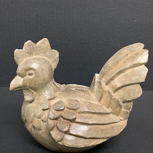 Puede incluir: Una figurita de pollo de madera tallada a mano con plumas detalladas y un pico realista. La figurita est&aacute; pintada en un color marr&oacute;n claro y tiene un aspecto r&uacute;stico.