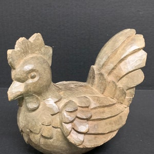 Puede incluir: Una figurita de gallo de madera tallada de color marr&oacute;n claro. El gallo est&aacute; de pie con las plumas de la cola extendidas.