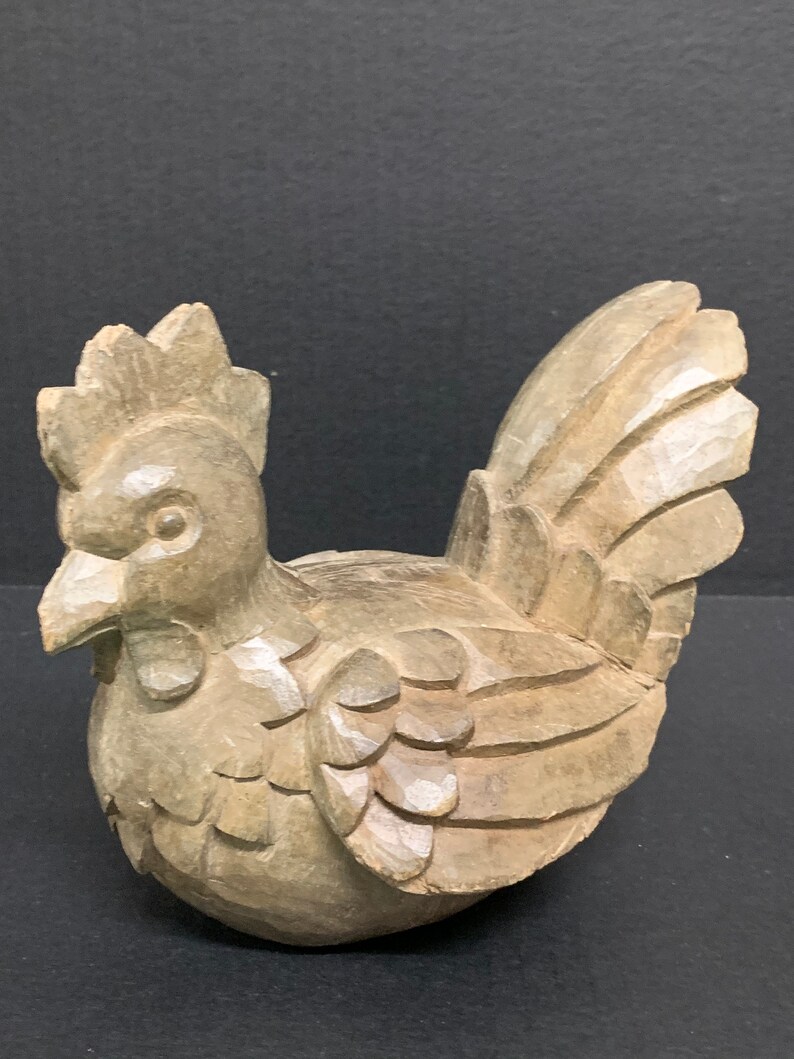 Puede incluir: Una figurita de gallo de madera tallada de color marr&oacute;n claro. El gallo est&aacute; de pie con las plumas de la cola extendidas.