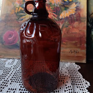 May include: A vintage brown glass jug with a handle and the word "PURE" embossed on the side. The jug has a textured pattern on the bottom and is likely used for storing liquids. The background features a painting with flowers.